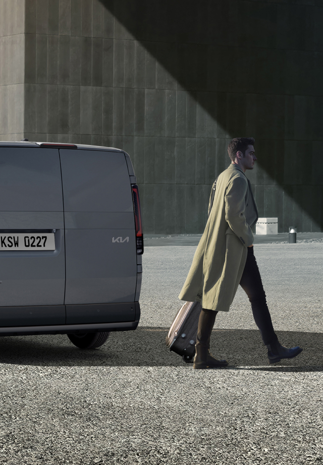 Dreiviertel-Rückansicht des Kia PV5 Passenger, geparkt in der Nähe eines Gebäudes, während ein Mann vorbeigeht.